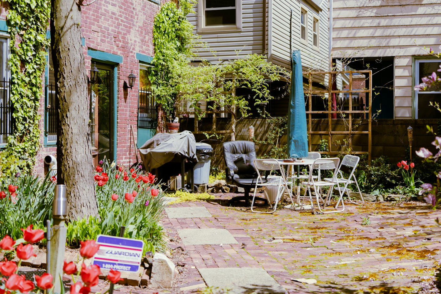 White patio furniture sits in a brick garden near two houses.